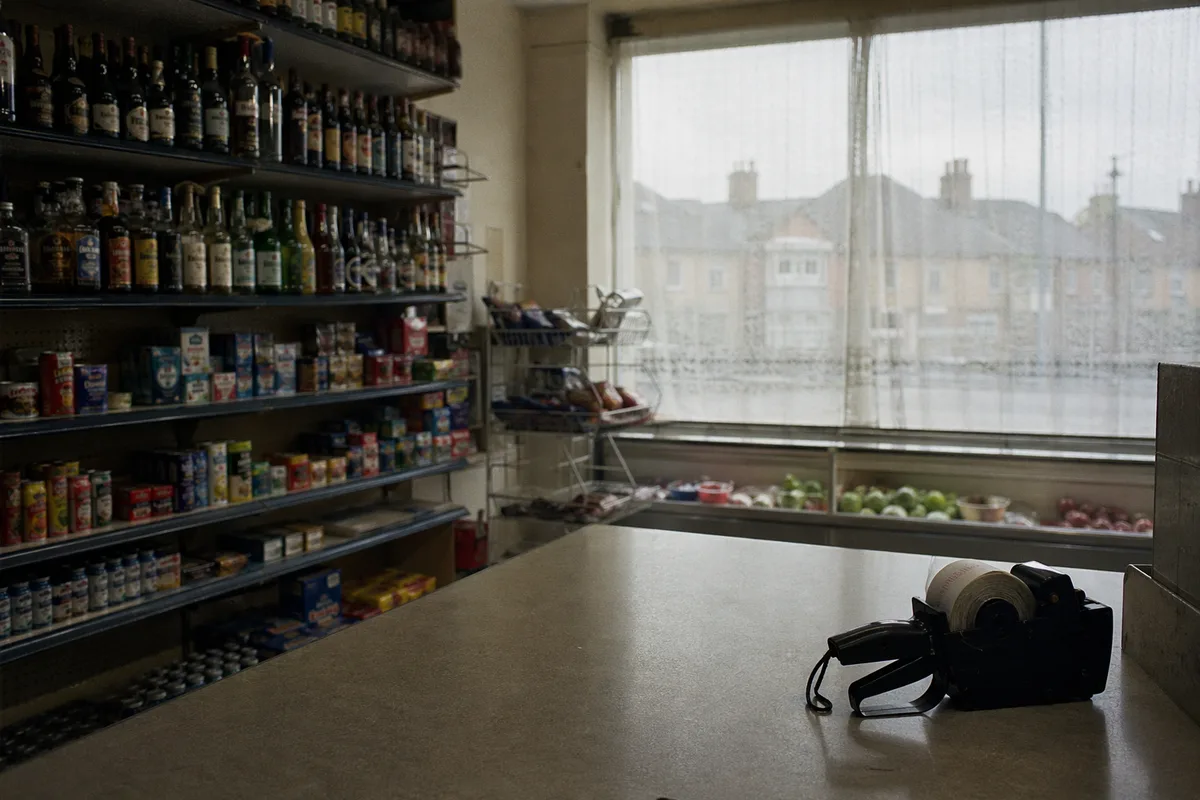 Empty British corner shop interior before opening hours, price gun on the counter, shelves of goods in the background.
